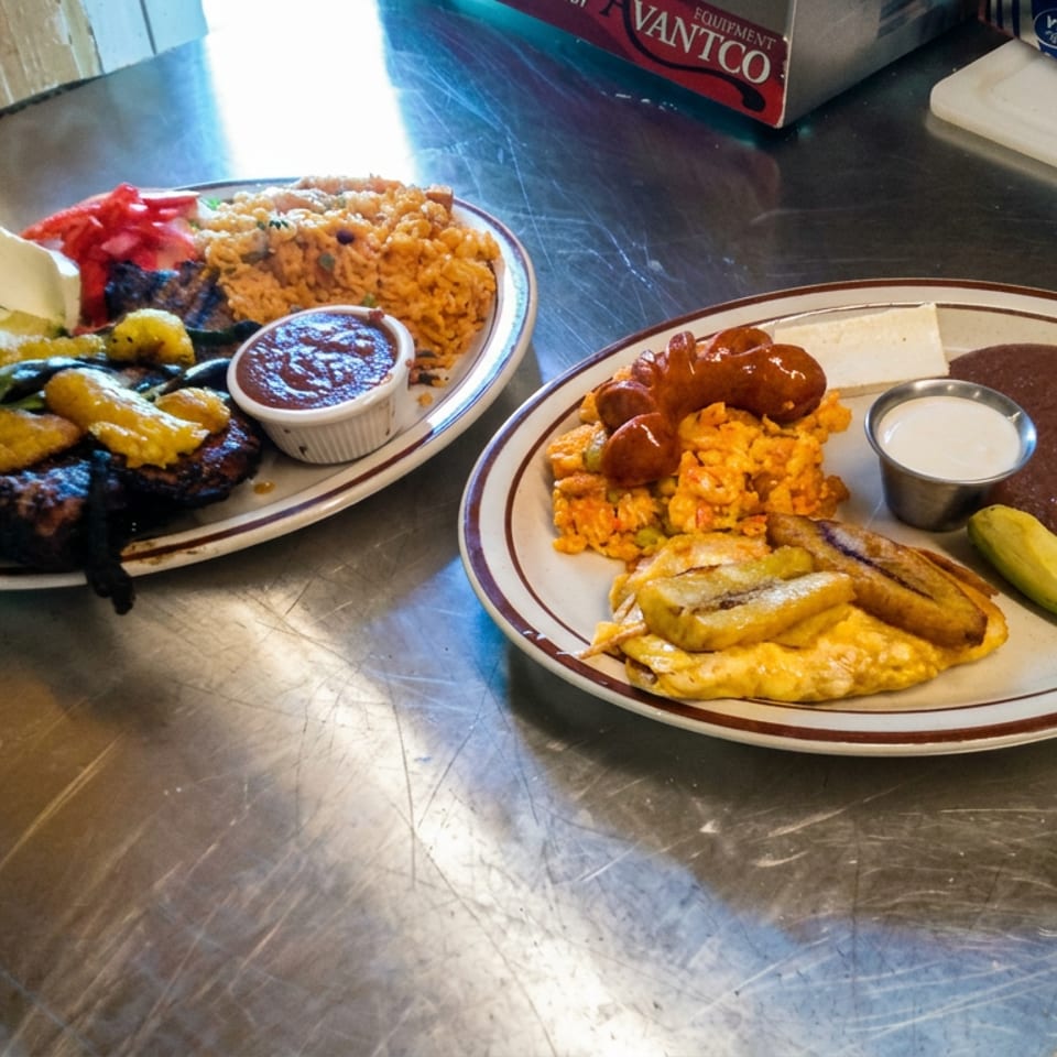 Two plates of Mexican food with grilled meats, rice, beans, enchiladas, avocado, lime, and sauces on a stainless-steel counter.